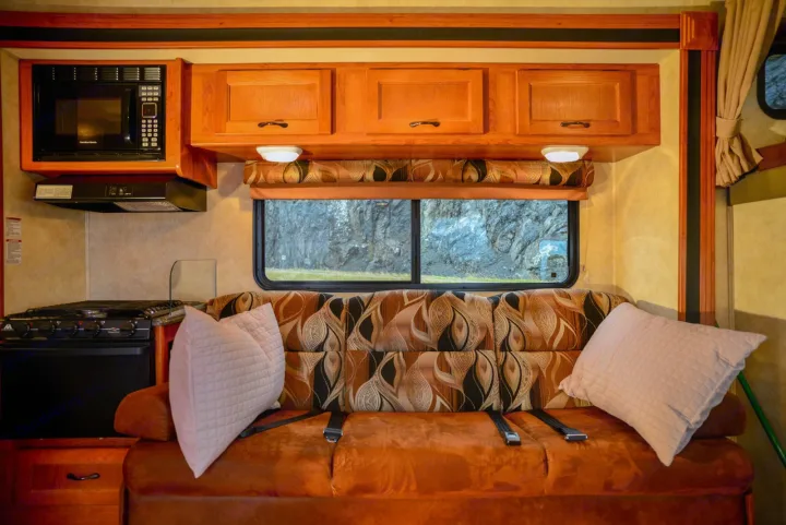 RV rental interior: compact kitchen appliances, cozy sofa with seatbelts, and a window framing a rocky mountain.. Forest River Coachmen Freelander 2014