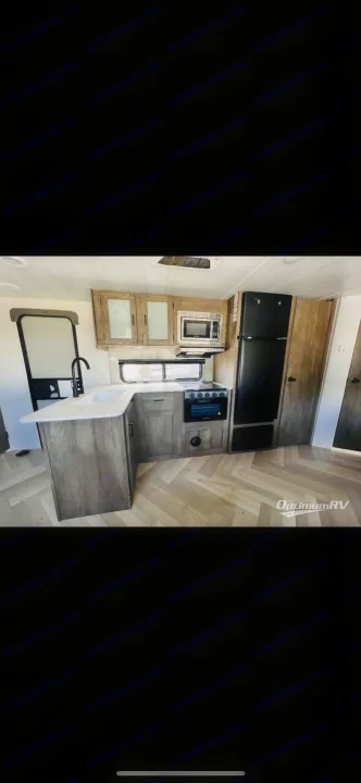 Modern RV rental kitchen with light wood cabinets, black appliances (fridge/stove), U-counter, sink, and herringbone flooring.. Forest River Wildwood X-Lite 2022