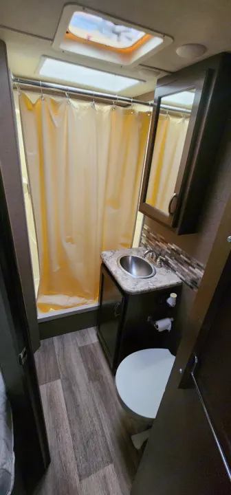Inside an RV rental bathroom: a yellow shower curtain, compact sink and vanity, and toilet with wood-look flooring.. Thor Motor Coach Ford E350 2018