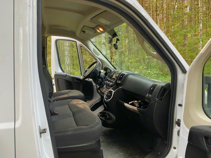 Clean white RV rental interior: front cabin, dashboard, and seats, with a beautiful forest scene visible through the windshield.. Ram Promaster 2018
