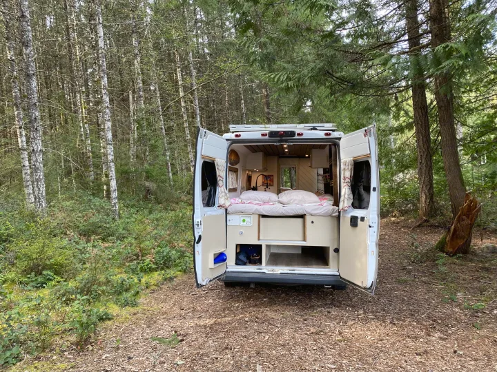 Open RV rental rear doors reveal a cozy bed & kitchen, parked on a forest path. Perfect for van life camping adventures.. Ram Promaster 2018