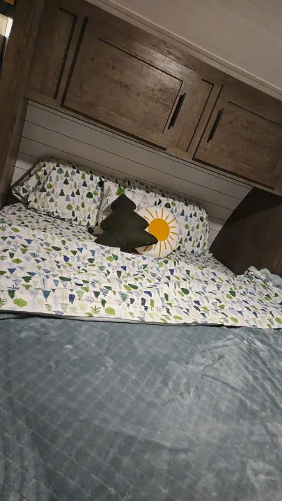 Cozy bed in an RV rental with tree-patterned bedding, a tree pillow, and a sun pillow, under wood cabinets & shiplap.. Forest River Salem 2021