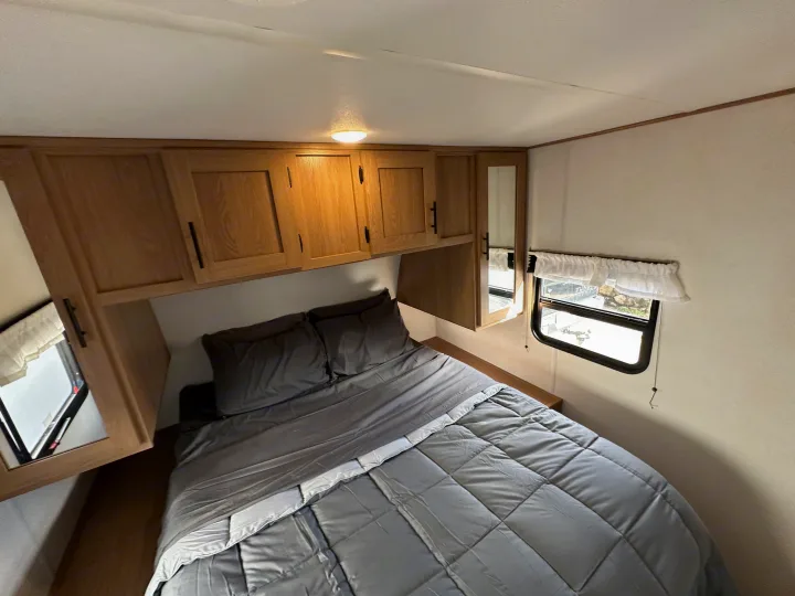 Interior of an RV rental bedroom featuring a bed with gray bedding, wooden overhead cabinets, and a window.. Fmc Primetime 2022