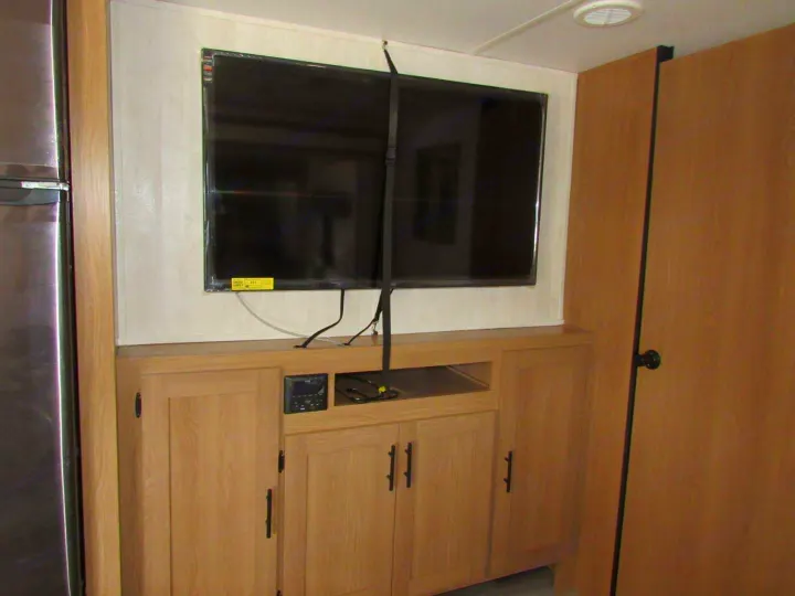 RV rental interior showing a wall-mounted TV secured with straps over a wood entertainment unit. Refrigerator and door are visible.. Fmc Primetime 2022
