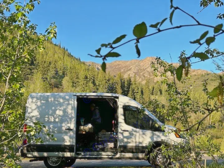 RV rental parked in a majestic mountain forest under clear skies, side door open for outdoor adventure preparation.. Ford Transit 2022
