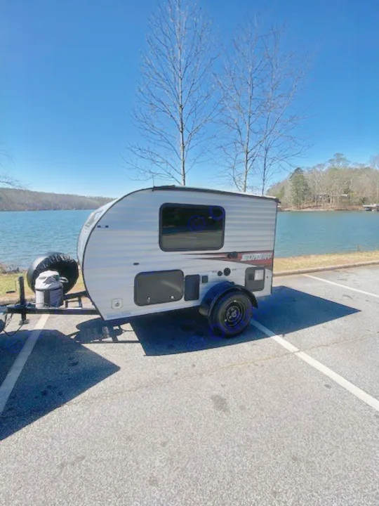 This compact RV rental, a small travel trailer, is parked by a scenic lake under a clear blue sky.. Sunset Park RV SunRay 2019