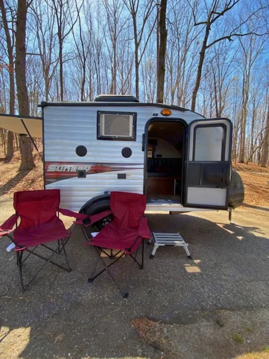 A small RV rental travel trailer is parked at a campsite, with two red chairs and a step, ready for camping.. Sunset Park RV SunRay 2019