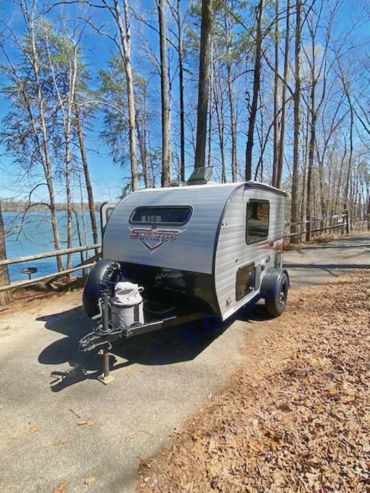 A compact RV rental is set up near a tranquil lake and trees, perfect for a peaceful camping getaway.. Sunset Park RV SunRay 2019