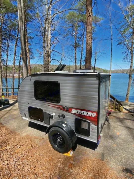 Sunny day RV rental at a scenic lakeside campsite. A gray travel trailer sits among trees by a blue lake, ideal for camping.. Sunset Park RV SunRay 2019