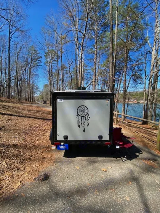 This compact RV rental, with a dreamcatcher decal, is perfectly parked by a lake, offering a serene outdoor escape.. Sunset Park RV SunRay 2019
