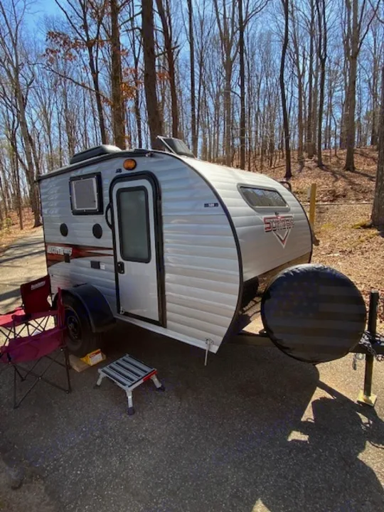 Silver teardrop rv rental travel trailer parked in a sunny wooded campground. Features a spare tire with an American flag cover.. Sunset Park RV SunRay 2019