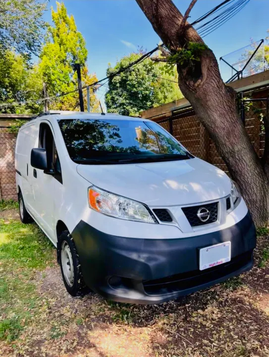 A white Nissan NV200 cargo van, ideal for an RV rental conversion, parked on grass beneath a large tree.. Nissan NV200 2015