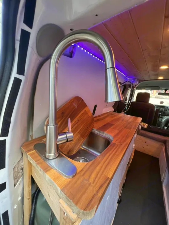 Inside a custom van conversion for RV rental: a compact kitchen setup with a sink, wood counter, and purple LED lights.. Nissan NV200 2015