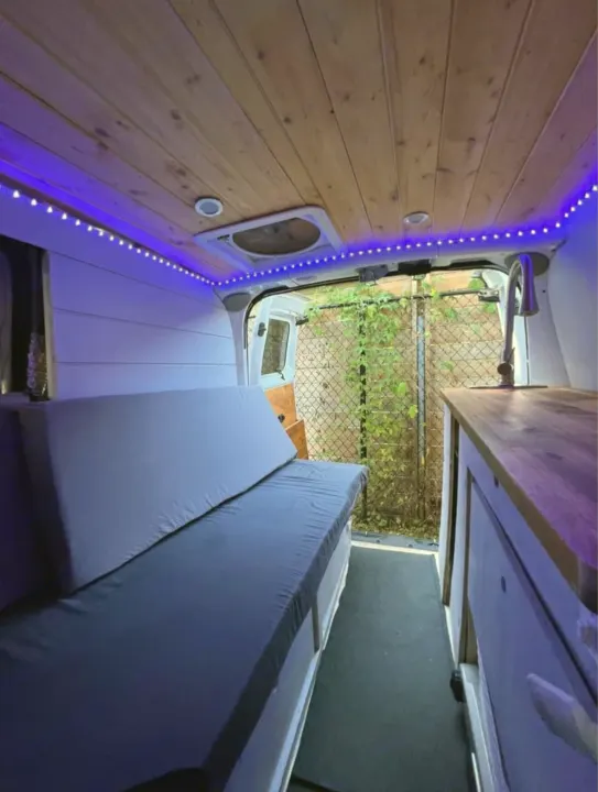Inside a custom RV rental: wooden ceiling, blue LEDs, comfy bed, and full kitchen setup visible.. Nissan NV200 2015