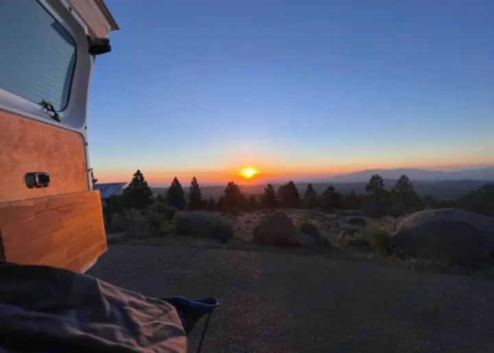 Golden hour peace from an RV rental. Open doors frame a sunset over distant mountains and pine trees.. Nissan NV200 2015