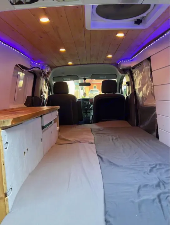 Inside an RV rental van: A bed fills the space, next to white storage and under a wood ceiling with warm lights.. Nissan NV200 2015