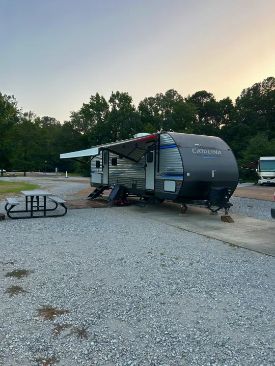 A Catalina RV rental travel trailer with an extended awning is parked at a gravel campsite beside a picnic table.. Forest River Coachmen Catalina Legacy 2020
