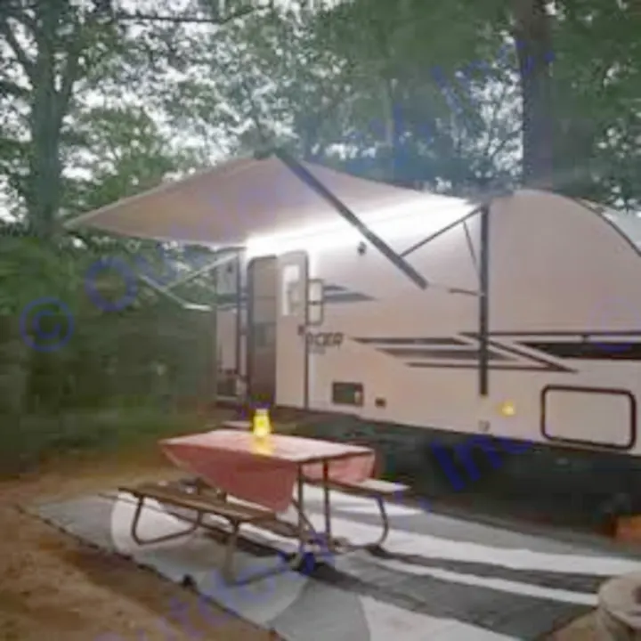 An RV rental camper at a lit evening campsite. Bright awning lights illuminate a picnic table, lantern, and mat.. Forest River Tracer Breeze 2020