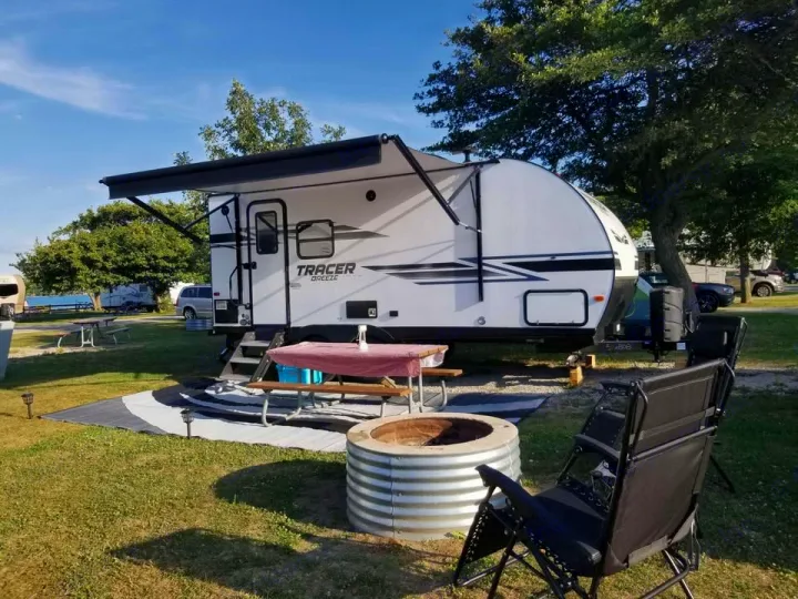 An RV rental is set up at a sunny campsite, featuring an extended awning, picnic table, fire pit, and chairs for outdoor relaxation.. Forest River Tracer Breeze 2020