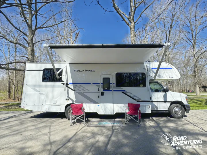 A white Four Winds RV rental with an extended awning, two red chairs, and a mat set up at a sunny park campsite.. Thor Motor Coach Four Winds 28Z 2026