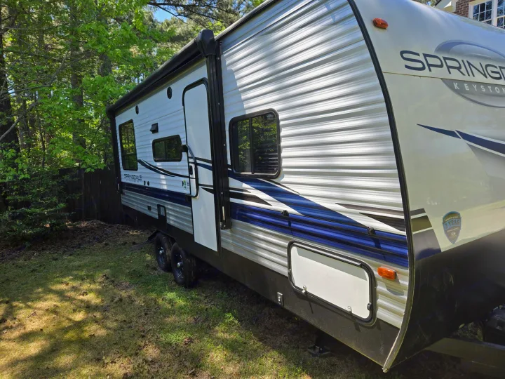 A white and blue Springdale travel trailer, perfect for an RV rental, sits on a sunny green lawn with trees.. Keystone RV Springdale 2023