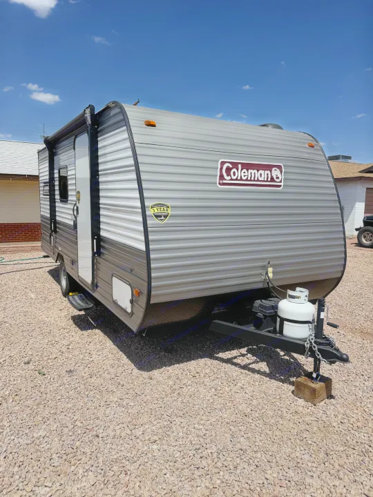 Grey Coleman travel trailer, ideal for RV rental. Parked on gravel, featuring an awning & front propane tank.. Dutchmen Coleman 17B 2024