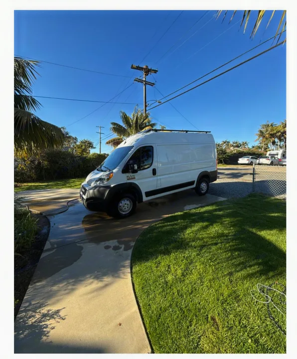 This white camper van, ready for RV rental adventures, is parked on a wet driveway on a sunny day.. Ram Promaster 2022