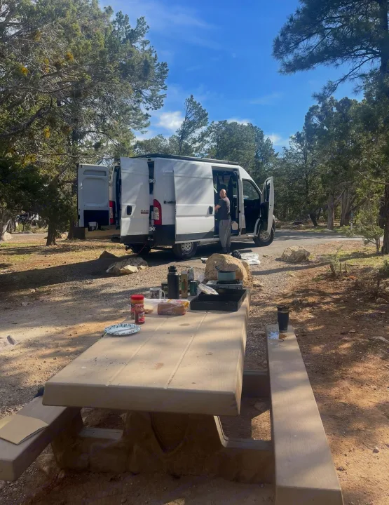 An RV rental camper van parked at a scenic campsite, with a person by the open van and a picnic table set for a meal.. Ram Promaster 2022