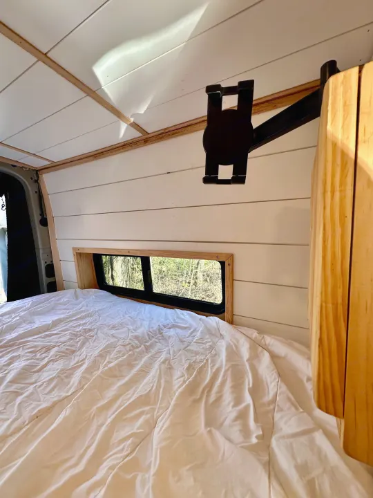 This RV rental's cozy sleeping area features a comfortable bed, white paneled walls, and a window with a tree view.. Mercedes-Benz Sprinter 2022