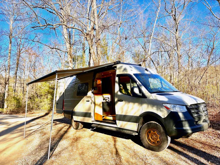 Explore nature! This custom camper van RV rental with an open door and awning, is parked in a wooded area on gravel.. Mercedes-Benz Sprinter 2022