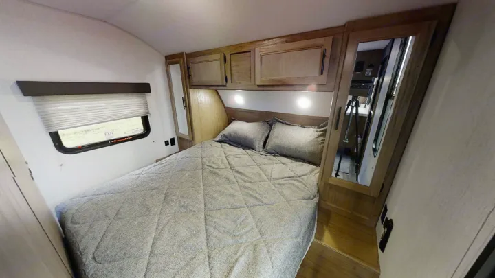 Compact RV rental bedroom showing a bed with gray bedding, overhead cabinets, and a window with blinds.. Forest River 26' Bunkhouse with Slide 2025