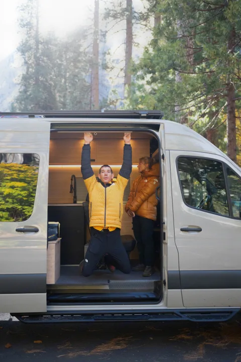 A man trains, hanging from an RV rental camper van doorframe, as a woman watches nearby. Outdoor setting.. Mercedes-Benz Sprinter 2024