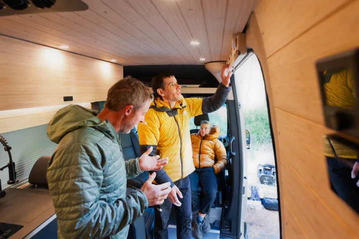 Inside this RV rental, two men in puffy jackets discuss its modern wood-paneled interior and features, with a woman seated.. Mercedes-Benz Sprinter 2024