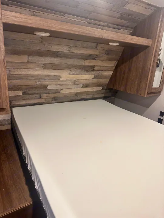 RV rental interior: a cozy bedroom with a clean mattress, rustic wood paneling, overhead shelf, and reading lights.. Crossroads RV Zinger 2020