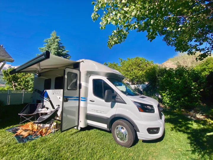 Sunny outdoor scene: a white camper van/rv rental on grass, awning out, with camping chairs & a dog relaxing.. Ford Transit 2023