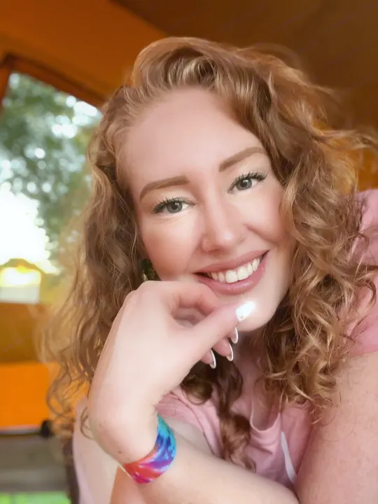 Smiling woman with curly red hair, wearing a pink top and tie-dye bracelet, happy in her RV rental.. Volkswagen Kombi T2 Westfalia 1976