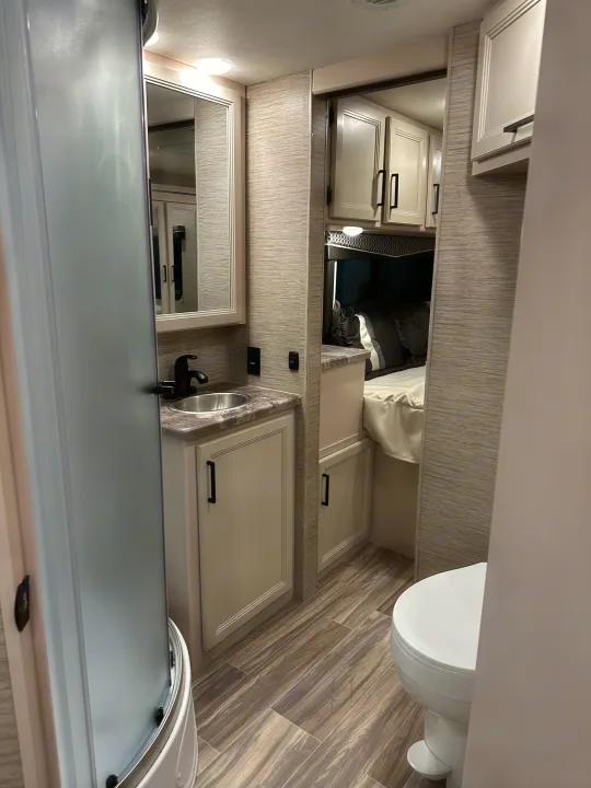 Modern RV rental interior with a compact bathroom featuring a frosted shower, sink vanity, toilet, and adjacent bedroom.. Coachmen Pursuit 2023