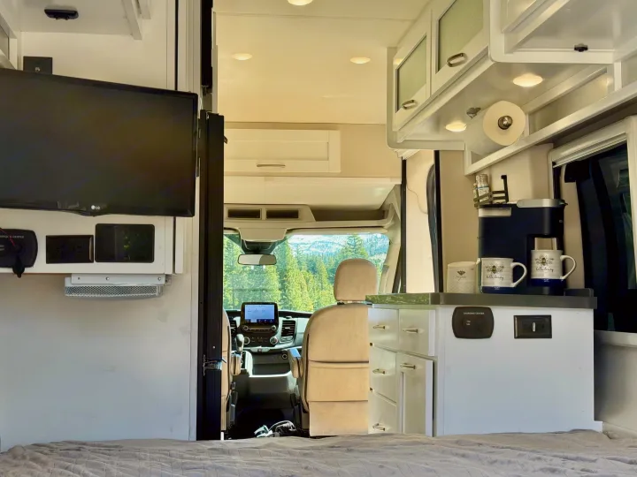 Inviting rv rental interior with a modern kitchenette, TV, and a stunning mountain forest view through the front cab, perfect for adventure.. Coachmen Beyond 2021