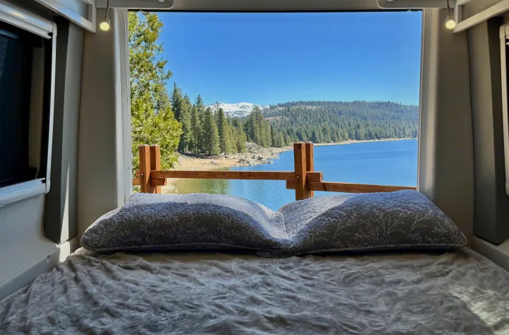 Serene RV rental view: a cozy bed faces a window overlooking a tranquil lake, lush forest, and distant snowy mountains.. Coachmen Beyond 2021