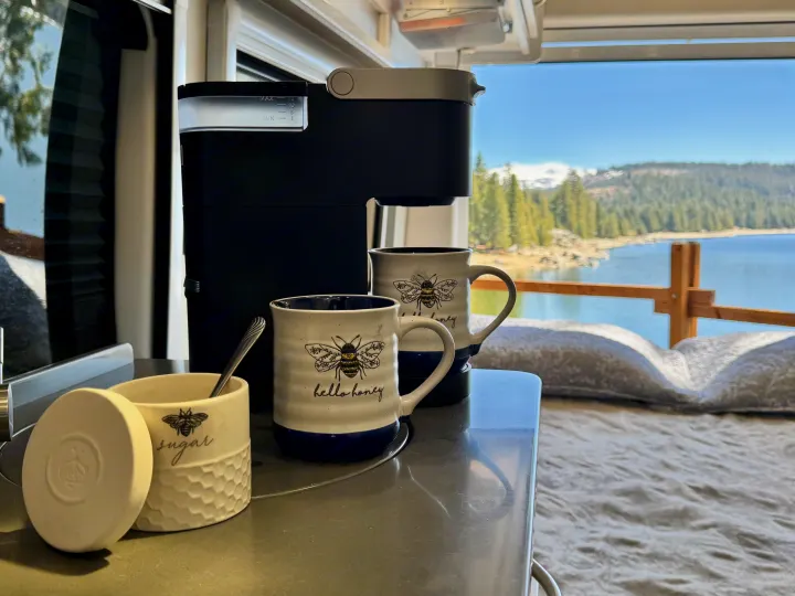 Coffee with a view! An RV rental interior showing a coffee maker, bee mugs, and sugar pot overlooking a scenic lake and mountains.. Coachmen Beyond 2021