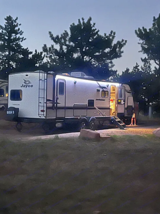Night view of a bright Jayco rv rental, a white travel trailer, parked at a campsite with trees. Person visible at steps.. Jayco Jay Feather 2021