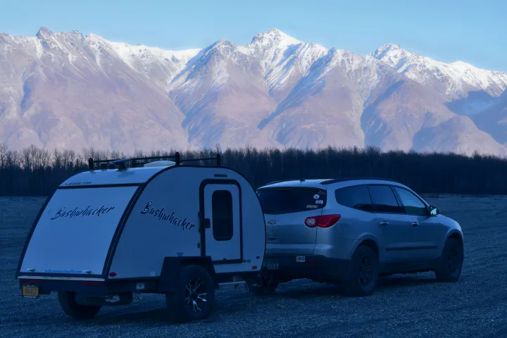 Bushwhacker RV rental and SUV on gravel, against majestic snow-capped mountains and bare trees.. Braxton Creek Bushwhacker 2022