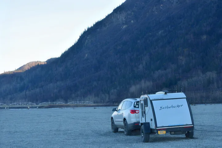 White SUV and Bushwhacker teardrop RV rental parked on gravel, with a mountain and bridge in the background.. Braxton Creek Bushwhacker 2022