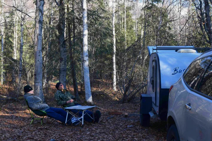 Two people relax in camping chairs by an RV rental and car in a peaceful forest setting.. Braxton Creek Bushwhacker 2022
