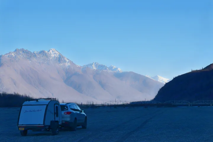 Explore scenic mountains with an RV rental! A teardrop trailer & SUV are parked on a gravel bar by bridges.. Braxton Creek Bushwhacker 2022