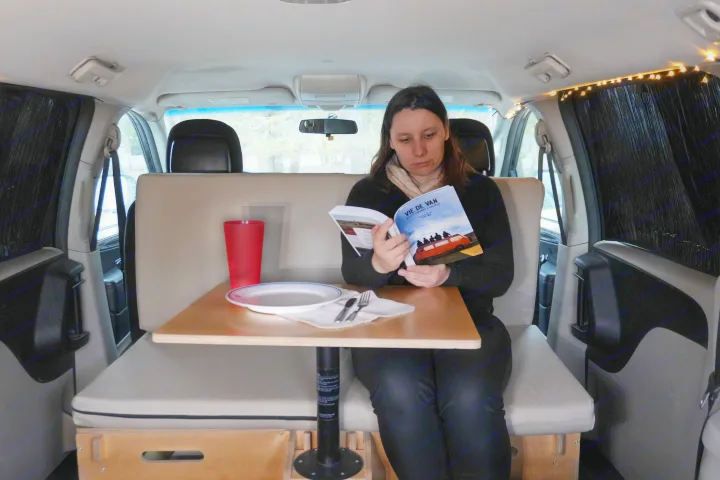 Woman reads "Vie de Van" in a cozy, converted RV rental. Shows interior with dining table, cushions, and string lights.. Dodge Grand Caravan 2014