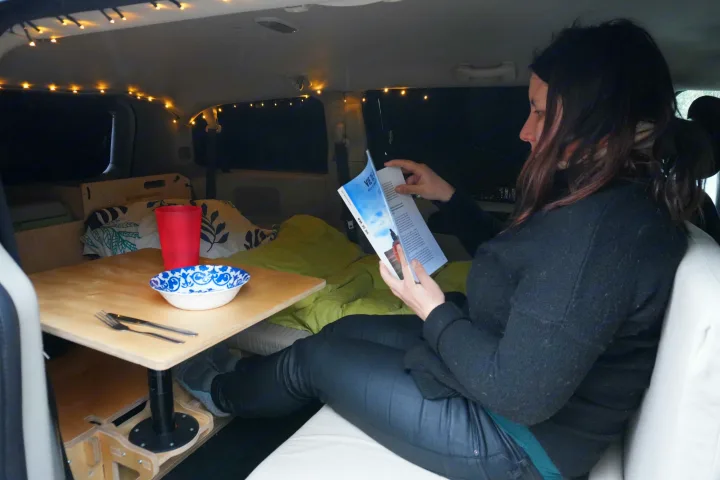 A person relaxes inside a cozy, customized RV rental, reading a book at a small table, illuminated by string lights.. Dodge Grand Caravan 2014