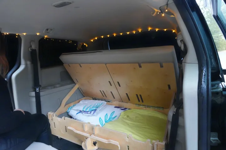 Cozy minivan camper conversion for an RV rental. Wooden bed frame with bedding & string lights inside.. Dodge Grand Caravan 2014