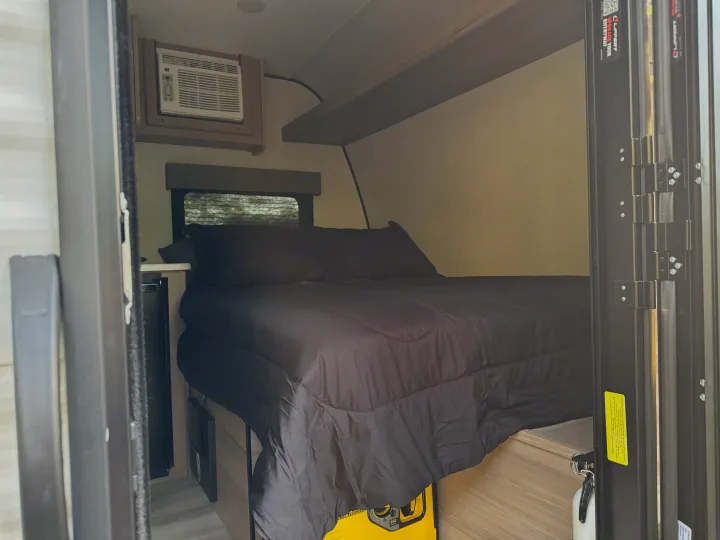 Cozy RV rental bedroom with black bedding, AC unit, window, and portable power. Compact and comfortable for travel.. Keystone RV Coleman 13B 2025