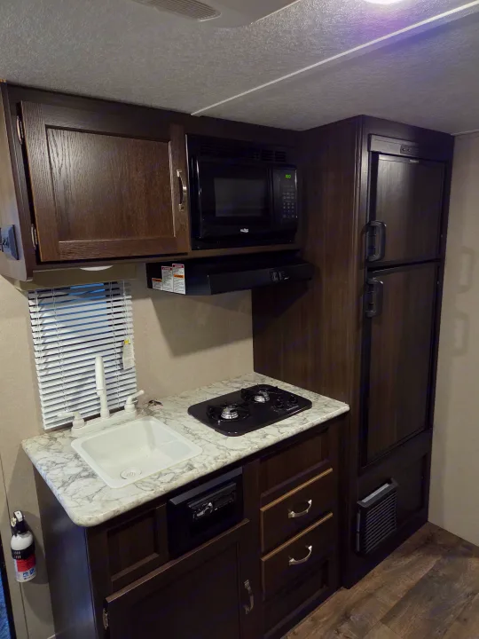 RV rental kitchen: Dark wood cabinets, white sink, stovetop, microwave, and refrigerator visible. A compact and well-equipped space.. Keystone RV Springdale 2019